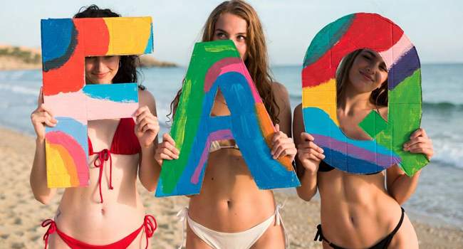 Tres estudiantes en la playa sostienen letras coloridas de FAQ durante un viaje de idiomas con LISA! en Malta