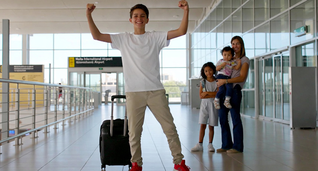 Llegada de un estudiante a Malta para un viaje de idiomas con los brazos levantados en pose de victoria con padres anfitriones al fondo
