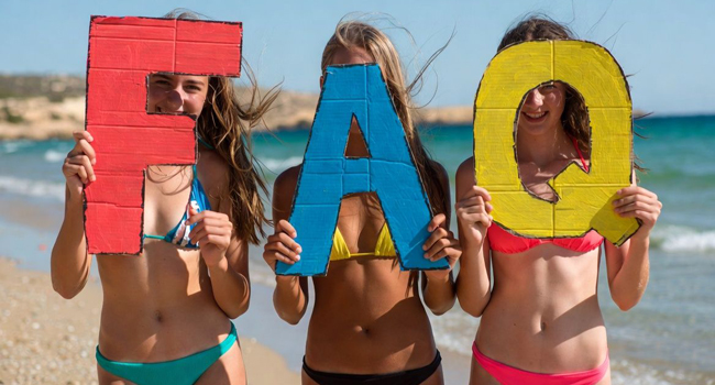 Viaje de idiomas para estudiantes en Malta: preguntas frecuentes para St. Julian’s Tres estudiantes de un viaje de idiomas para estudiantes en Malta en la playa de St. Julian’s con grandes letras FAQ